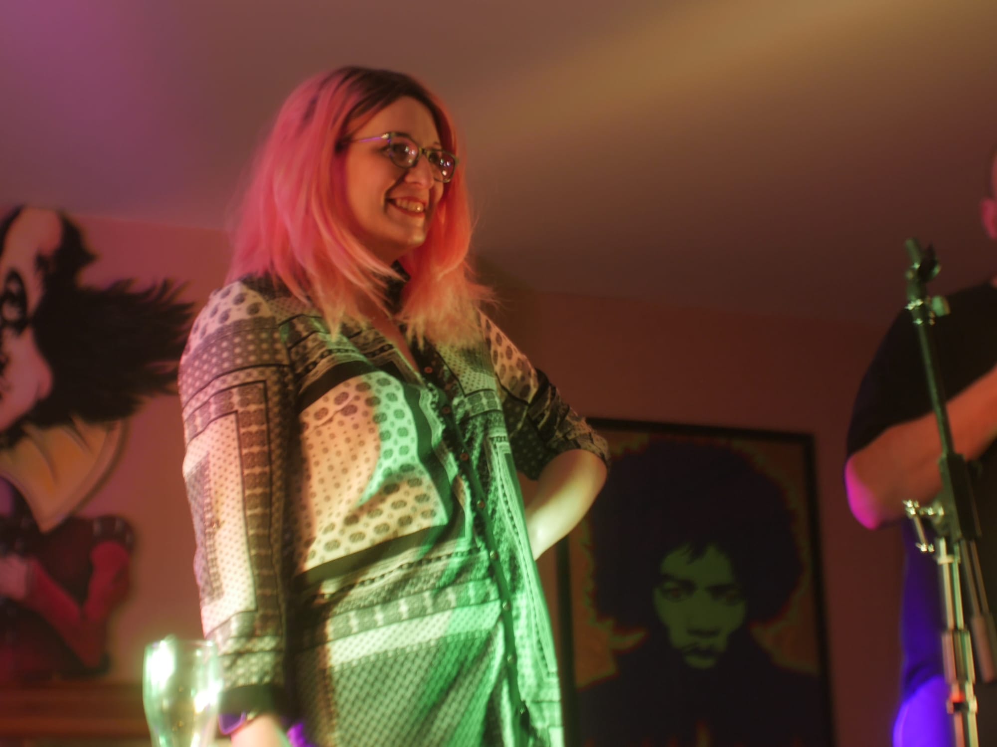 A photograph of Ava smiling on stage at a comedy show. She has long pink hair and glasses. There is a mic stand to the right of the shot