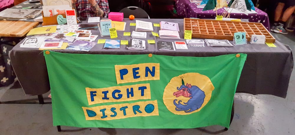 A table laid out with a few zines and prints, there's also a letterpress tray filled with badges. In the front there's a green/yellow/blue fabric banner, which says pen fight distro, next to a picture of a unicorn with a pencil for a horn