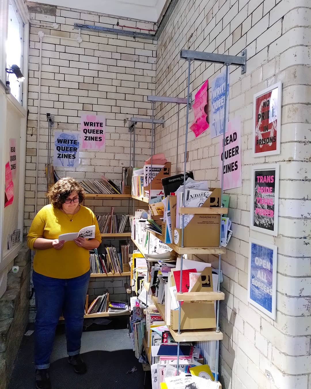 A small room lined with shelves that are full of zines. A white, brown-curly-haired person stands in the middle of the room reading a zine.