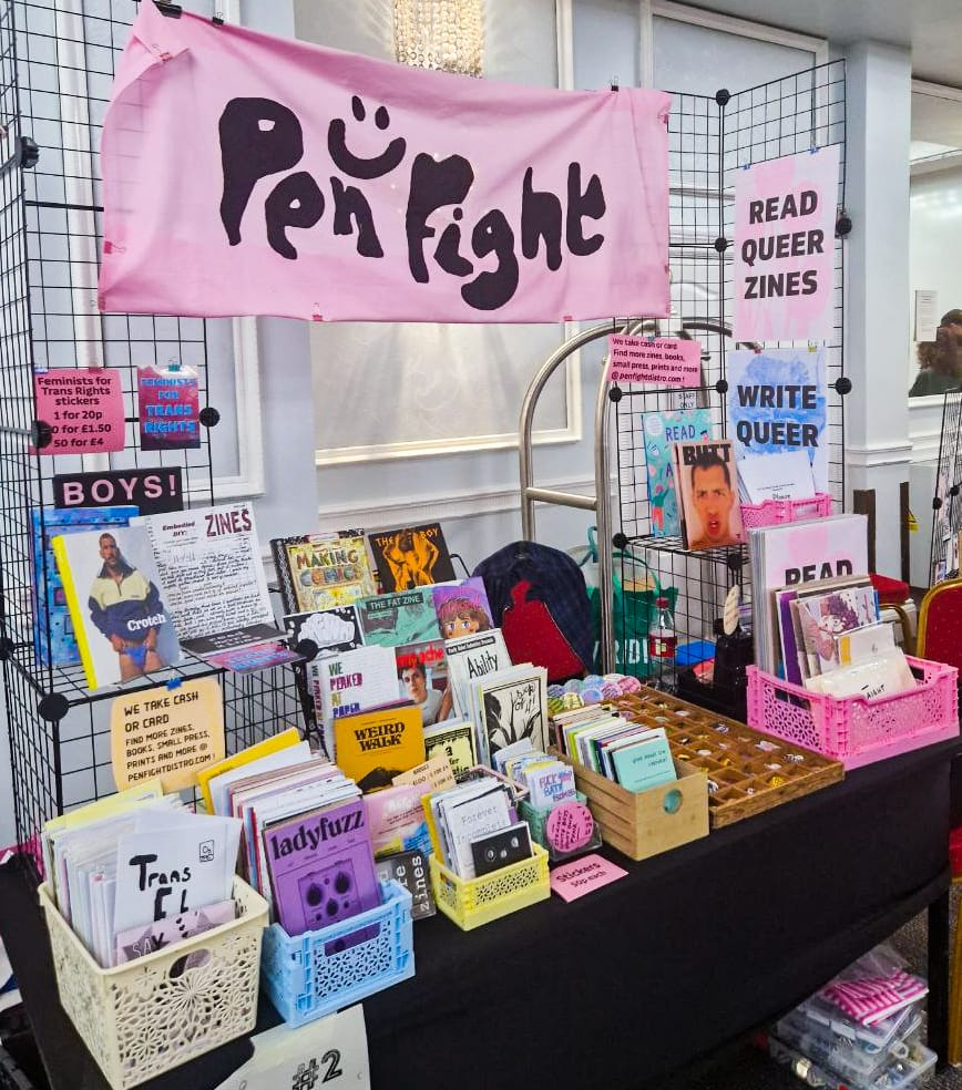 a table set up for selling things, there are layered racks absolutely full of different zines, plus a box of prints and a tray of badges. Above it all is a pink banner that says "Pen Fight" in black lettering