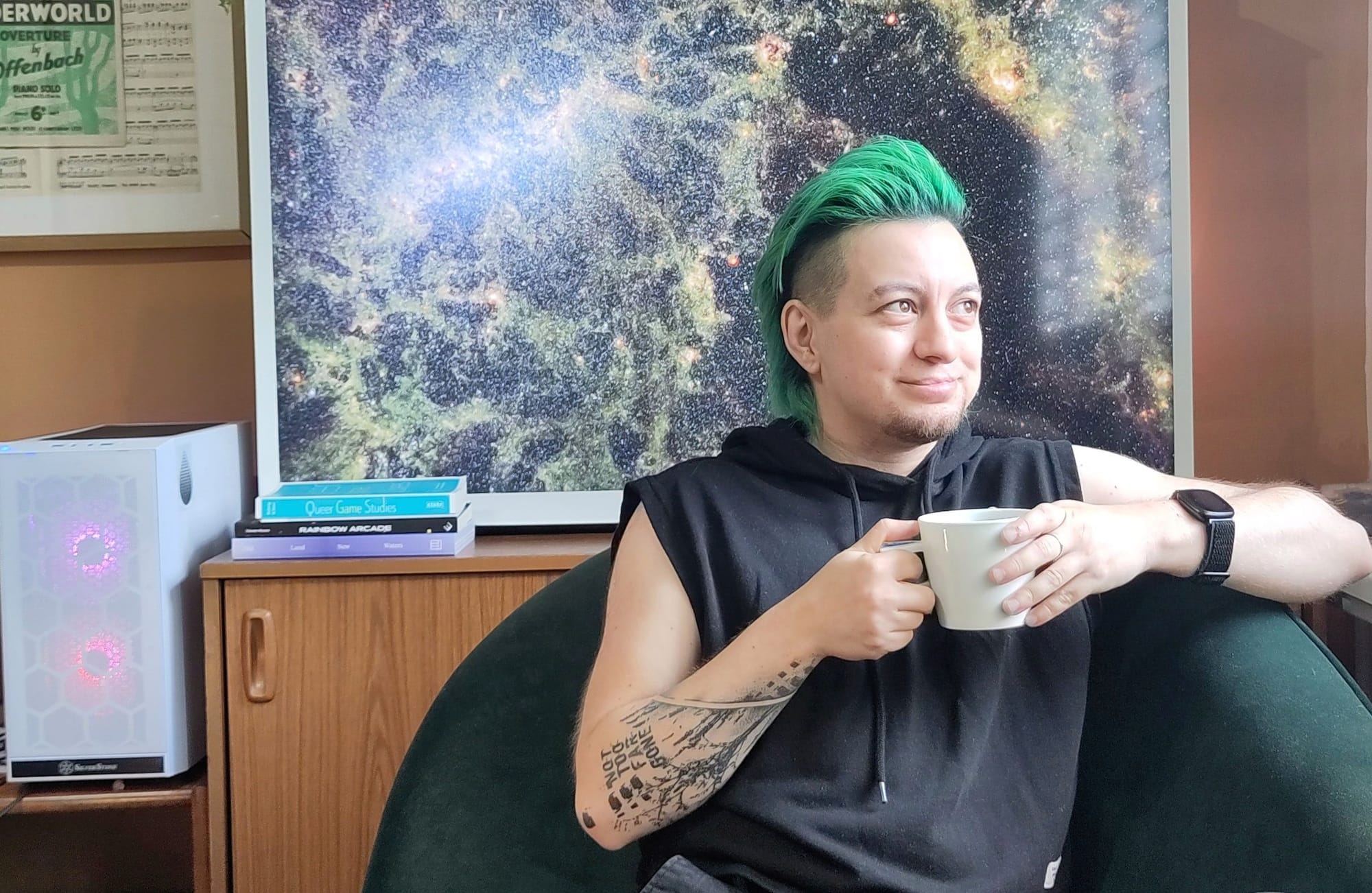 Photo of Zoyander Street: a small white trans man with green hair, sat on a green velvet chaise longue holding a porcelain mug in front of one of the James Webb Space Telescope images