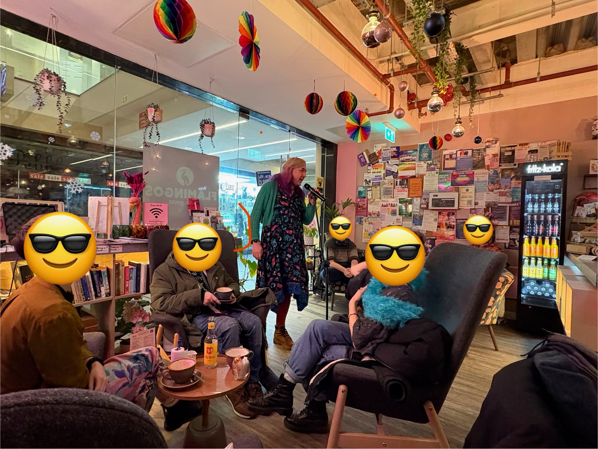A nonbinary person in a cardigan and floral dress addresses a small group in a coffee shop. There are various colourful mobiles and a noticeboard in the background.