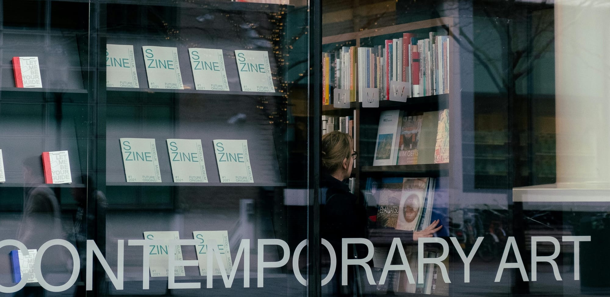 the window display of an art-book shop. Several zines labelled 'S Zine' are on shelves, along with a wider selection of books. The words 'contemperary art' are written on the window in capital letters.