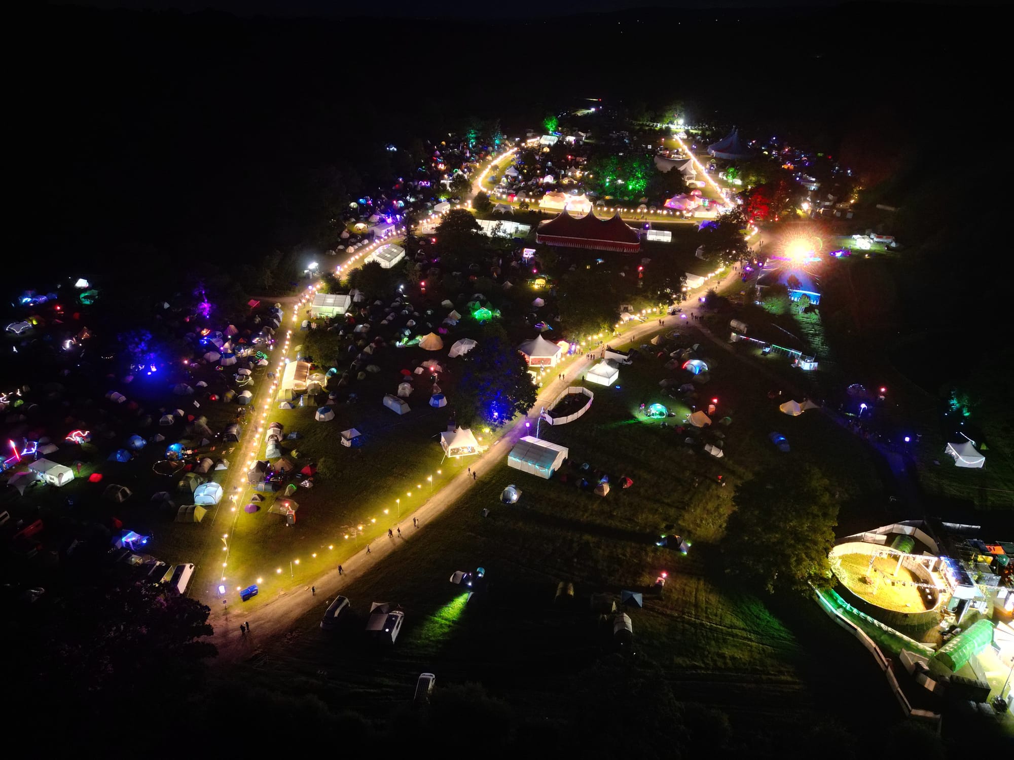 Alt text: an aerial view of the Electromagnetic Field site at night. It is very brightly lit with yellow-lit paths between many tents