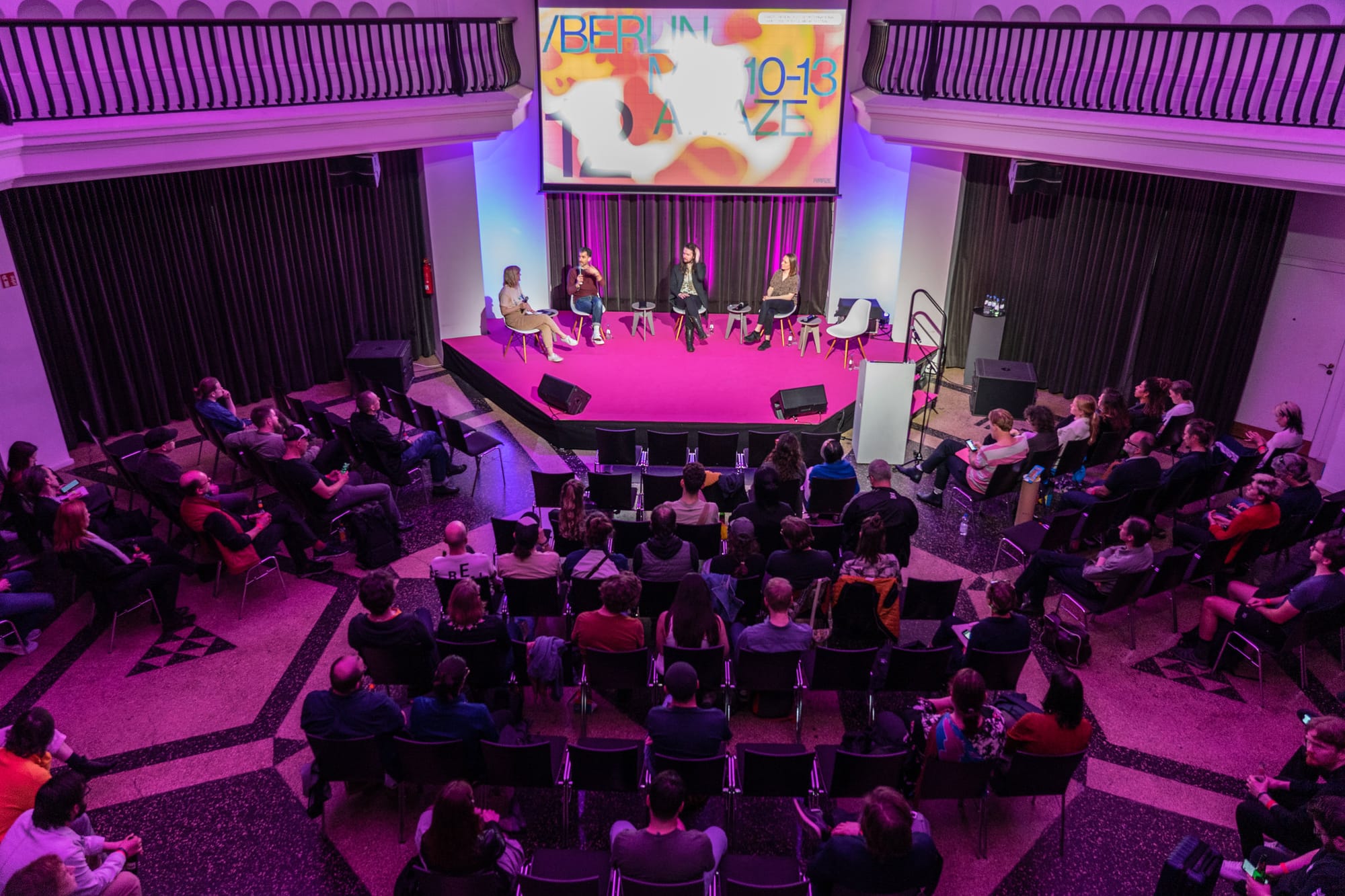 An audience bathed in pink light around a hexagonal stage. On the stage, four people sit in chairs, giving a talk. Behind them, a screen reads Berlin, May 10-13, A MAZE