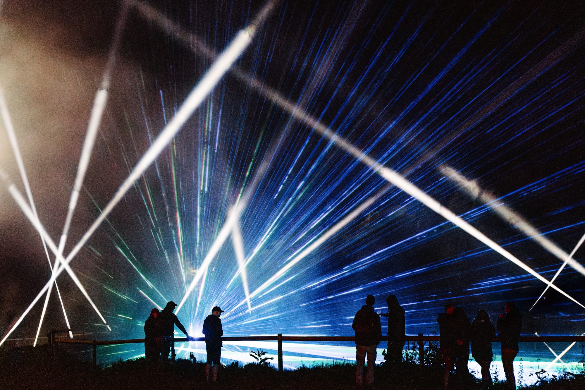 A night-photo of people silhouetted by a laser-light show.