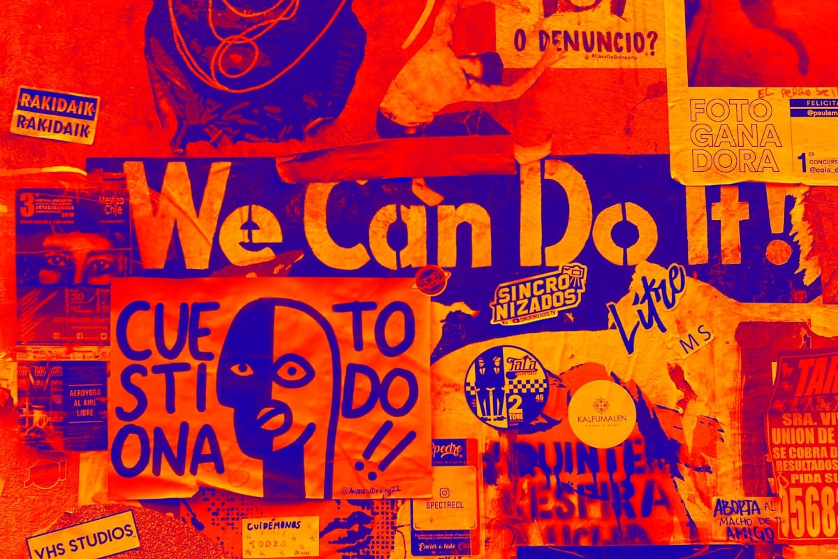 A colourful grafittied wall, covered with signs and posters in red and blue, with one saying 'We can do it!' and others in Spanish.