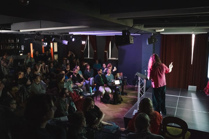 An audience is gathered around a stage at a small bar. Kim is on stage, who is a white transfemme with long hair, wearing a pink blouse with her back to us.