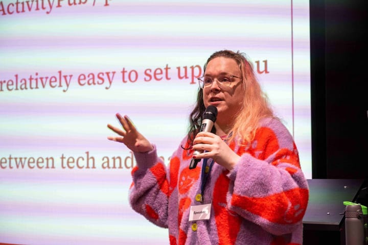 Kim, wearing a natty pink and orange cardigan and speaking into a microphone while giving a presentation. Behind her, a large set of presentation slides are visible.