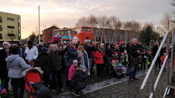 A group of people at Hulme Winter festival watching something off to a stage right of shot. There's various trees and a bus in the background, push chairs and people taking photos