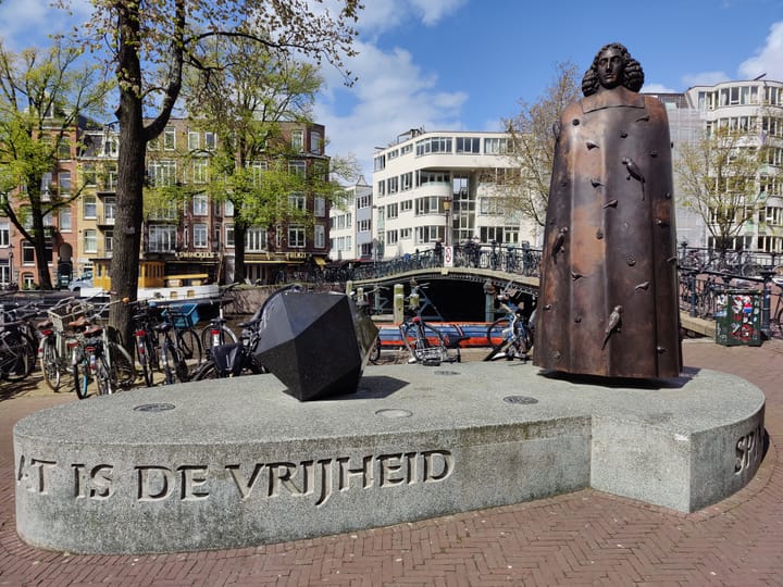 A large dark grey metal statue of Spinoza covered in birds on a concrete plinth. It has a dodecahedron on it. In the background is an Amsterdam street scene on a sunny d