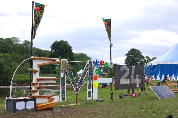 Alt text: The EMF 24 sign. Each letter is decorated differently, 'E' has a bamboo water flume, 'M' is covered in circuit boards, 'F' has pinwheel fans & the '24' are cut from black material.